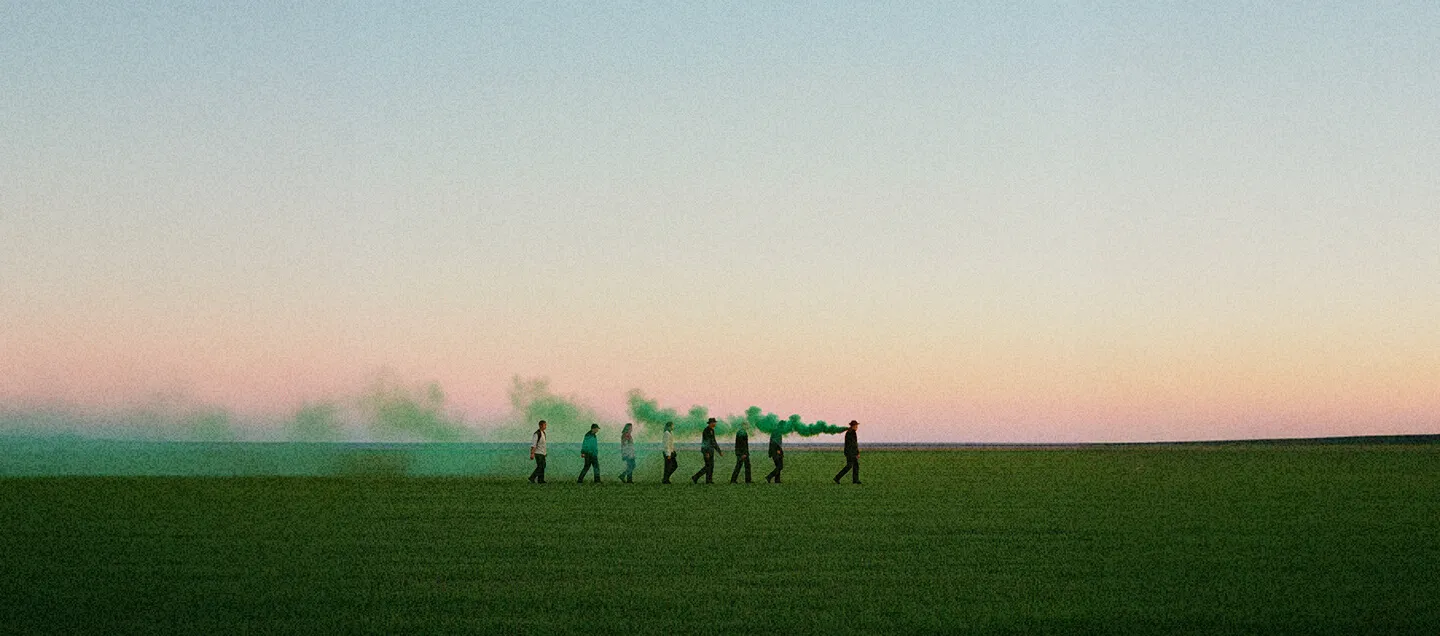 Sept personnes marchant en file indienne dans un champ vert avec une fumée verte s'échappant de la cinquième personne.