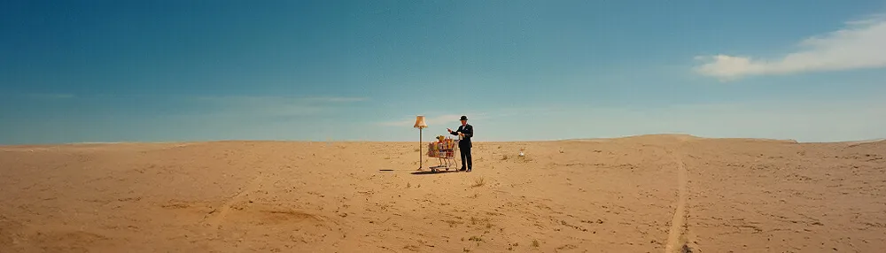 Homme en costume debout dans un désert avec un chariot rempli d'objets et une lampe sur pied à côté.