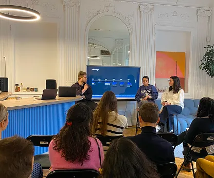 Discussion en groupe dans une pièce avec trois personnes assises devant un écran et plusieurs personnes écoutant en rangées de chaises.