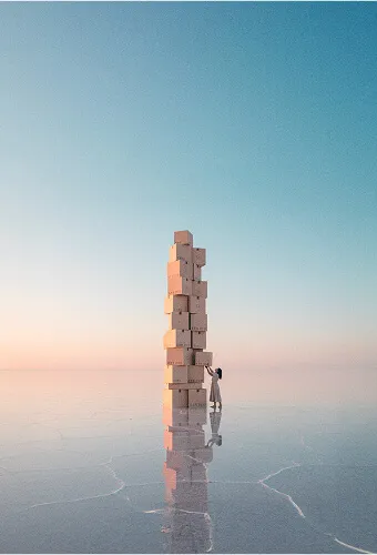 Personne empilant une très haute tour de cartons sur une surface réfléchissante sous un ciel clair au coucher du soleil.