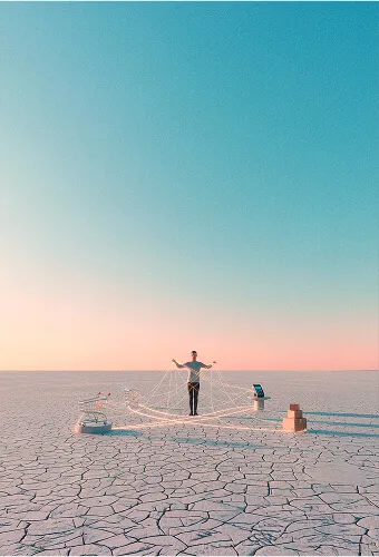 Personne debout sur un sol craquelé tendant les bras autour de fils connectés à des objets sur un fond de ciel bleu dégradé au coucher du soleil.
