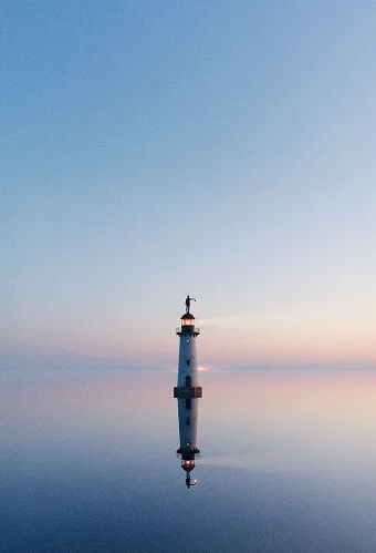 Phare blanc se reflétant dans une mer calme au coucher du soleil sous un ciel clair.