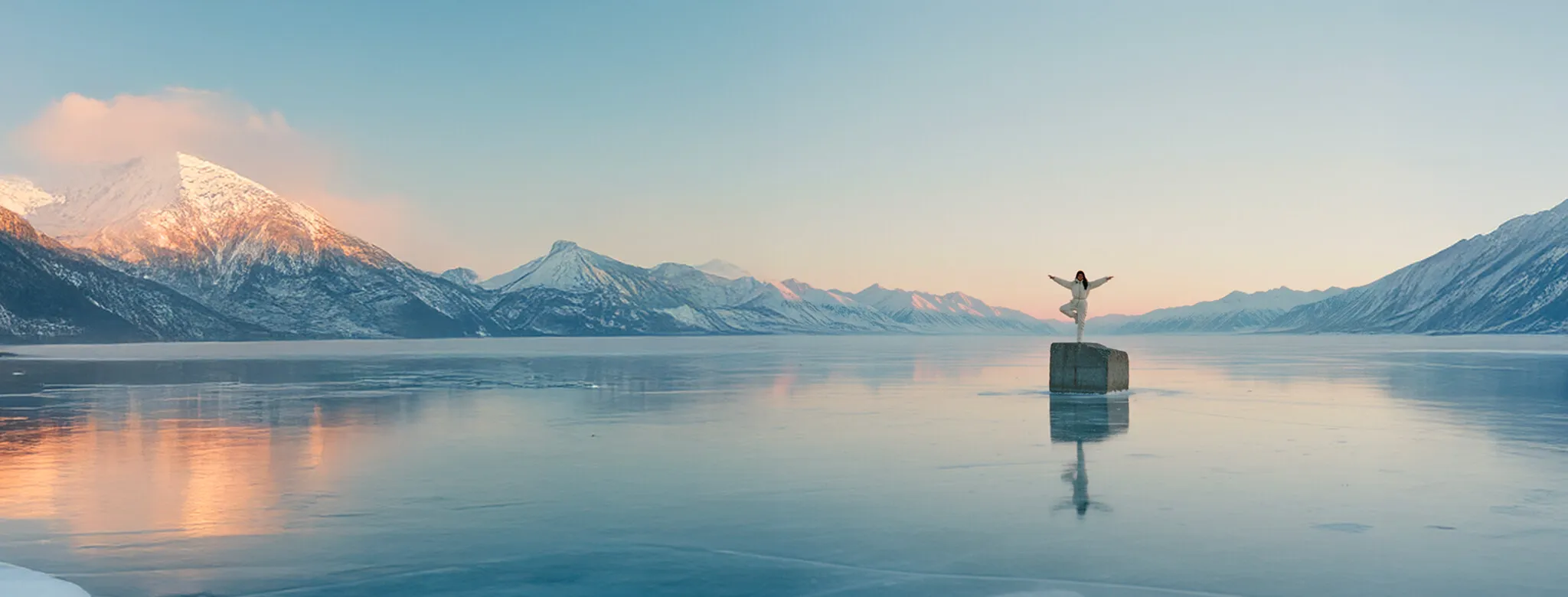 Person in winter clothes standing balanced on a rock in the middle of a frozen lake surrounded by snowy mountains at sunset.