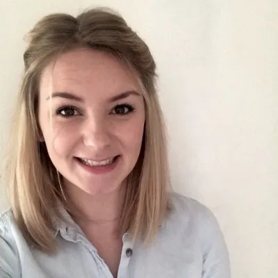 Jeune femme blonde souriante avec une chemise bleu clair devant un fond blanc.