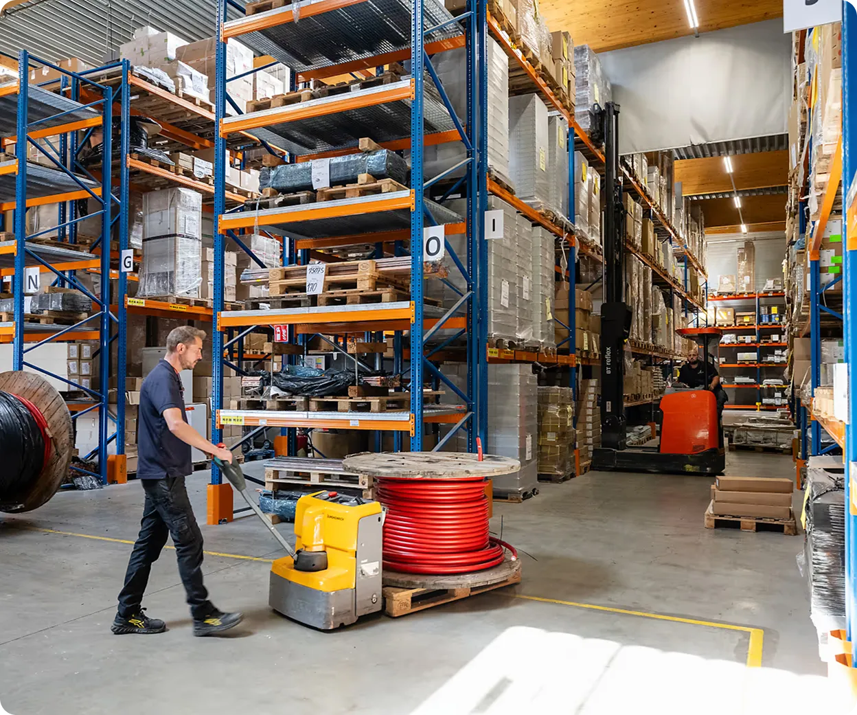 Un homme pousse un chariot élévateur électrique jaune transportant un rouleau de câble rouge dans un grand entrepôt avec des étagères remplies de cartons et palettes.