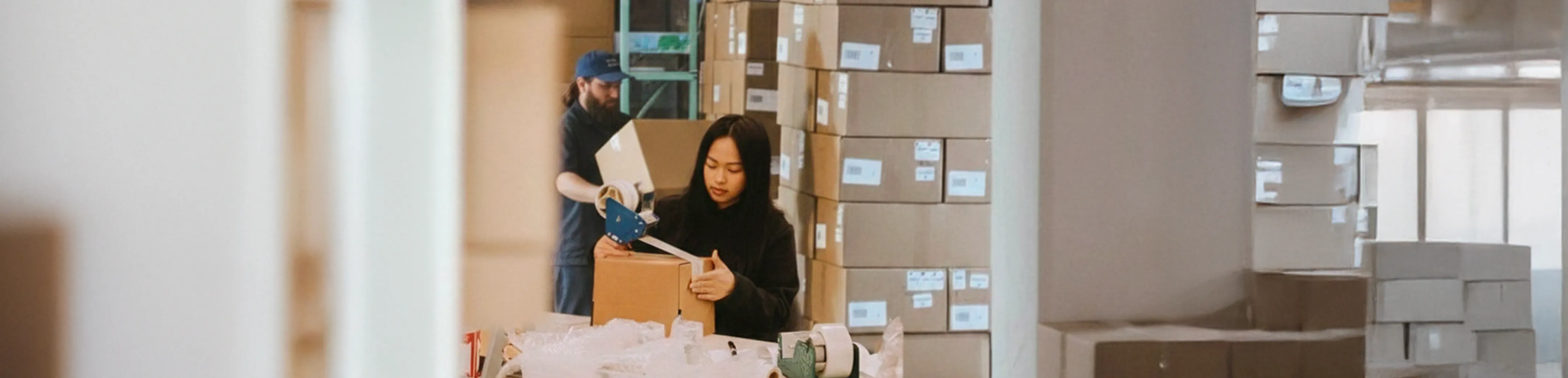 Deux personnes emballant et scellant des cartons dans un entrepôt rempli de piles de boîtes en carton.