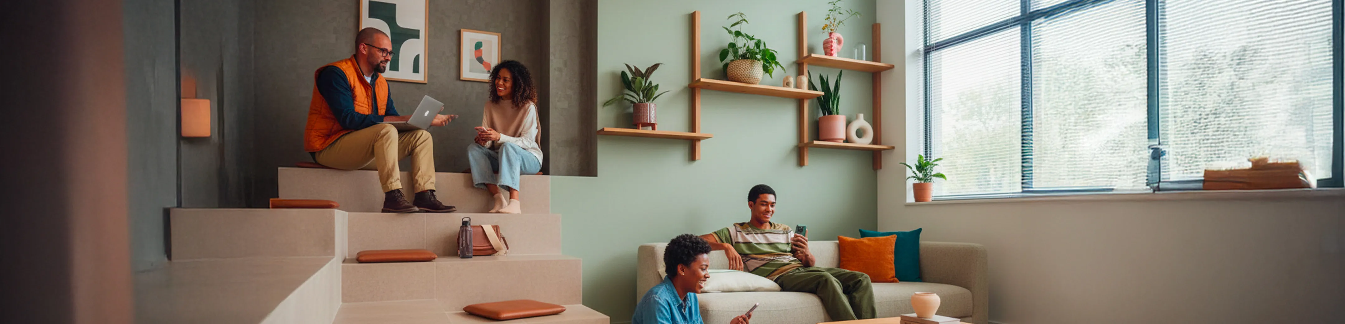 Quatre personnes décontractées collaborant dans un espace de travail moderne avec des fenêtres grandes, des étagères murales et des coussins orange.