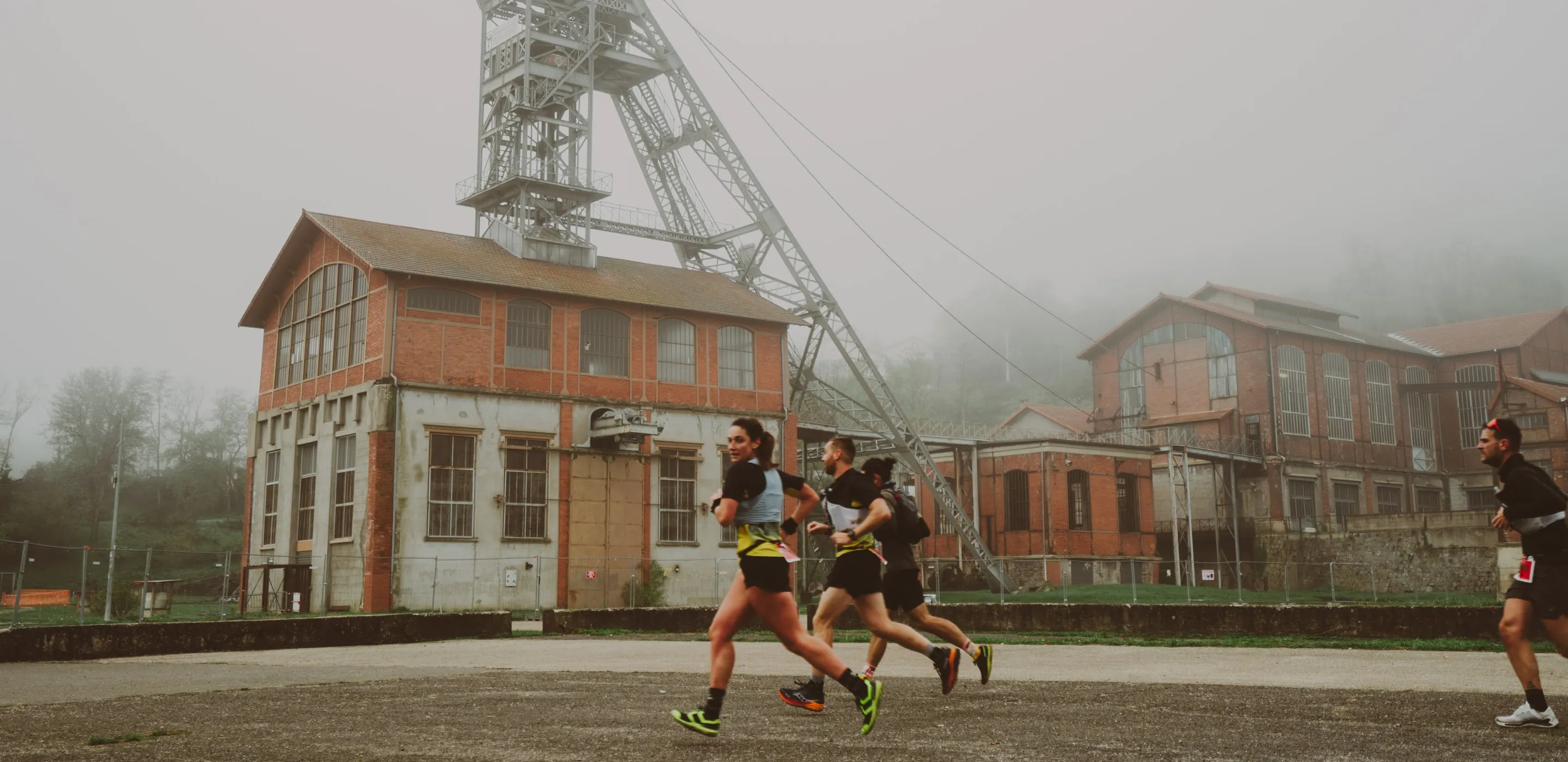 Quatre coureurs en tenue sportive courant devant un bâtiment industriel en brique avec une structure métallique élevée par temps brumeux.
