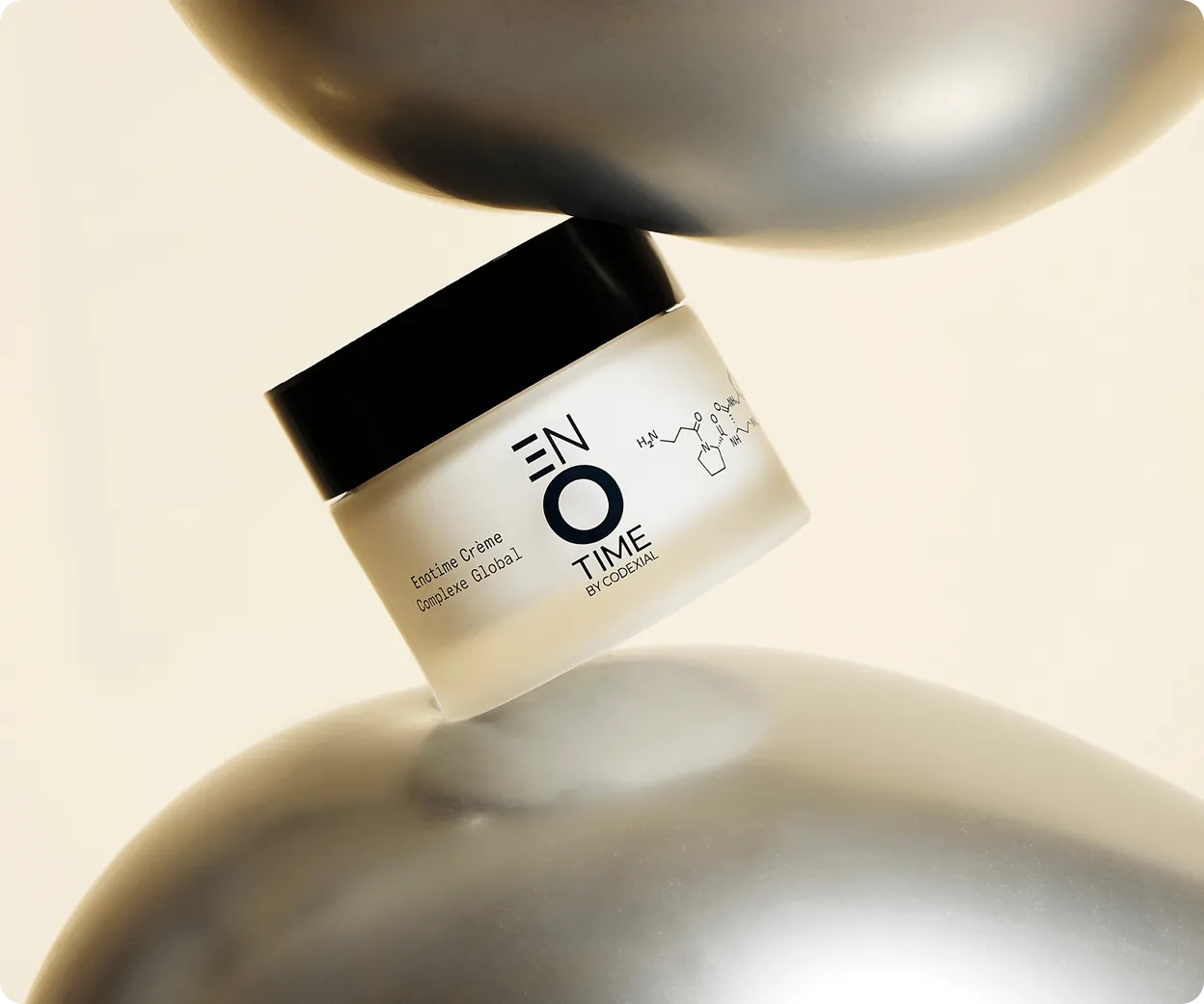 Pot de crème Enotime Complexe Global avec couvercle noir posé entre deux objets métalliques argentés arrondis.