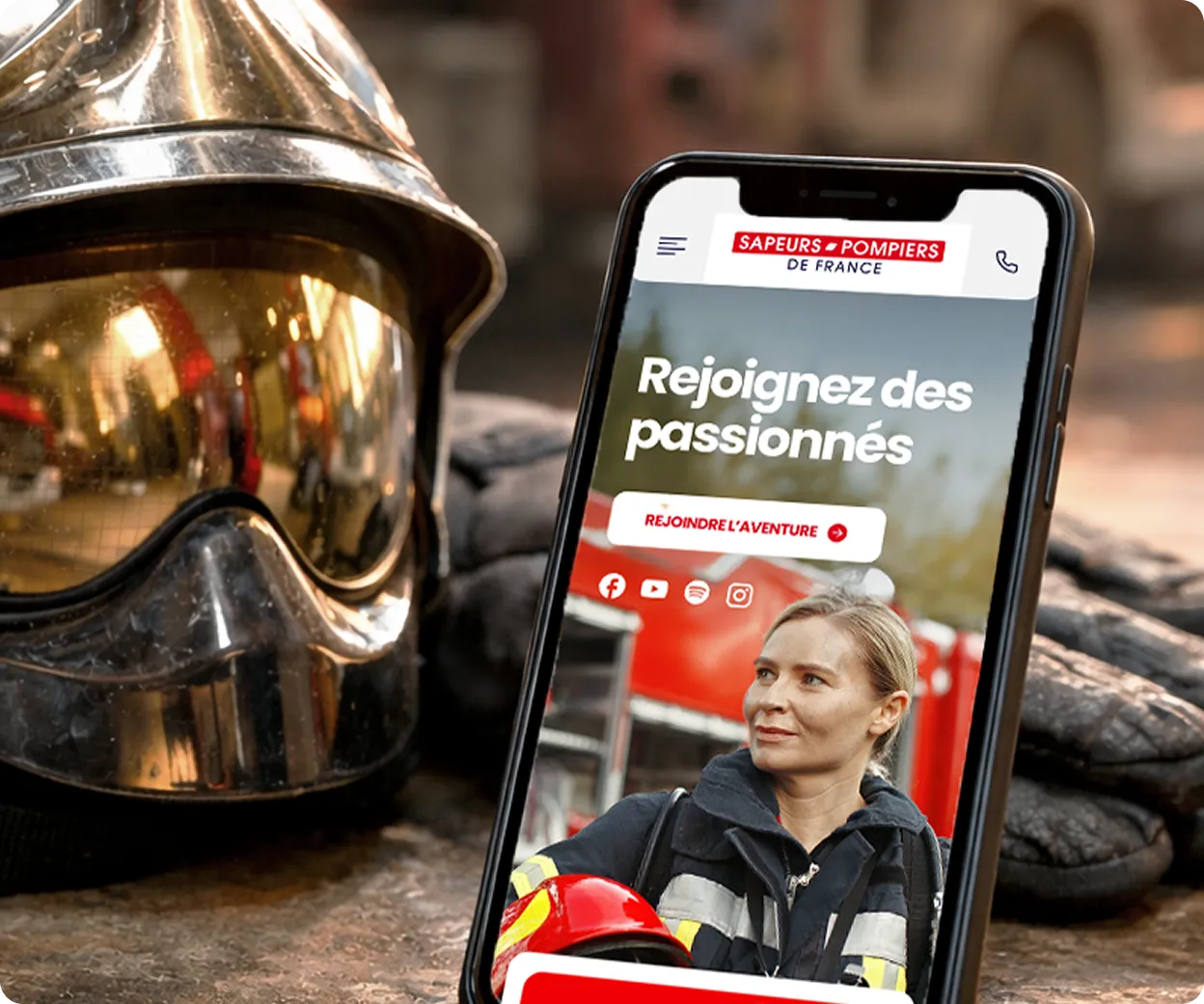 Un casque de pompier métallique et des gants posés à côté d’un smartphone affichant la page d’accueil des Sapeurs-Pompiers de France avec une femme pompier en uniforme.