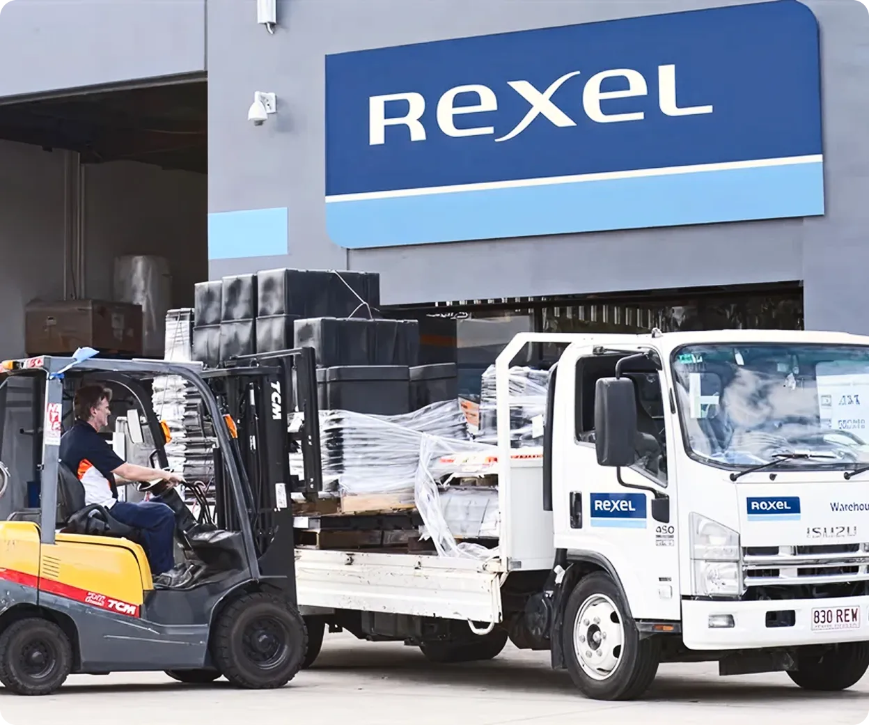 Un homme conduit un chariot élévateur qui charge des palettes emballées sur un camion blanc avec le logo Rexel devant un entrepôt Rexel.