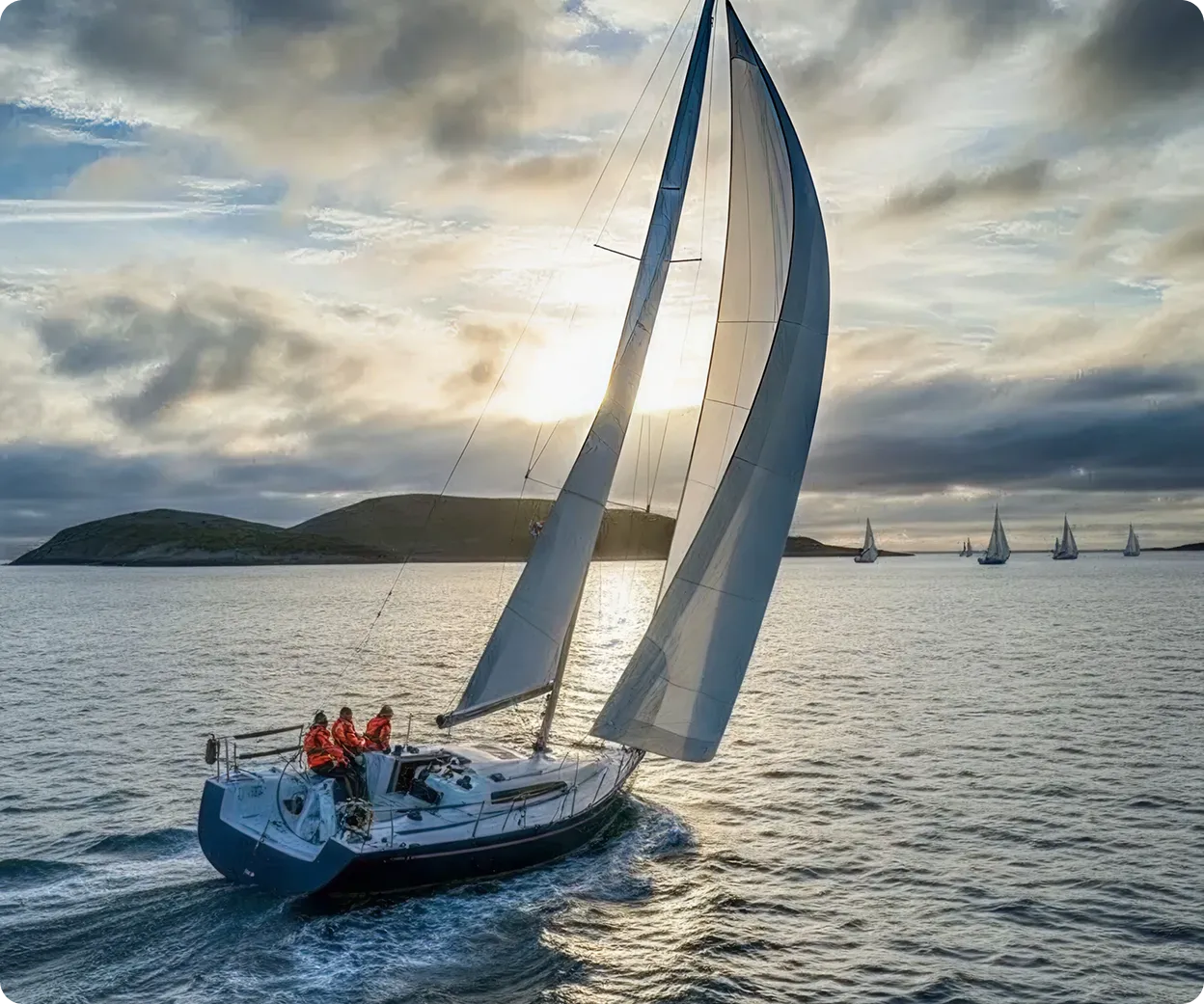 Un voilier avec trois personnes en vestes rouges navigue sur une mer calme au coucher du soleil, avec plusieurs autres voiliers à l'arrière-plan.