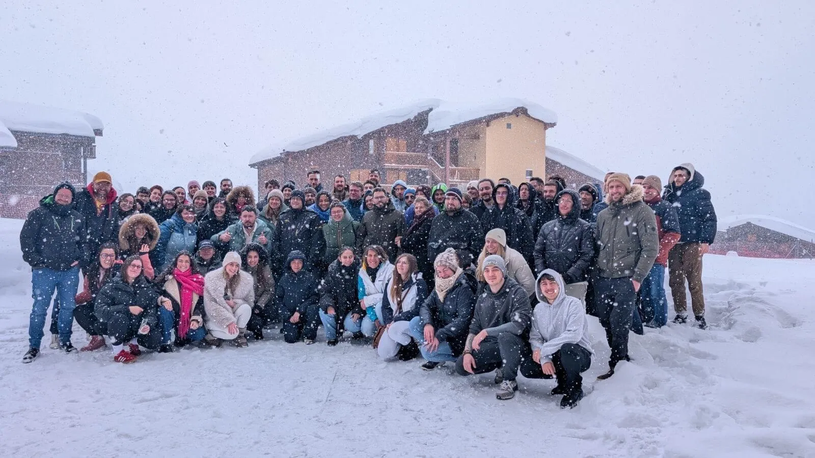 Groupe de personnes posant dans la neige devant des chalets en bois sous une légère chute de neige.
