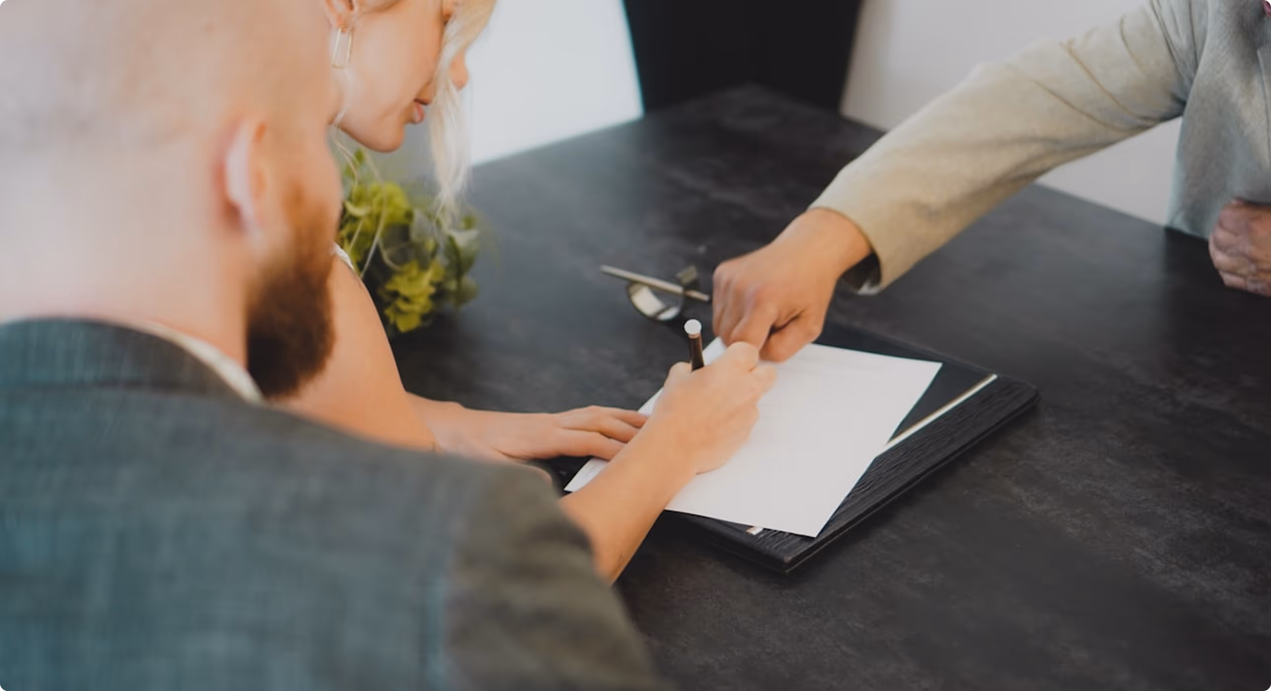 Person signing a document on a clipboard while another person points at the paper, and a third person watches.