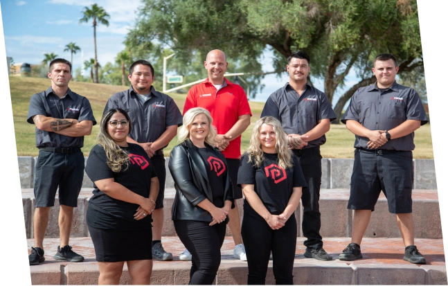 Group of eight people standing outdoors on steps with greenery and palm trees in the background, some wearing black shirts with a red logo and one person in a red polo shirt.
