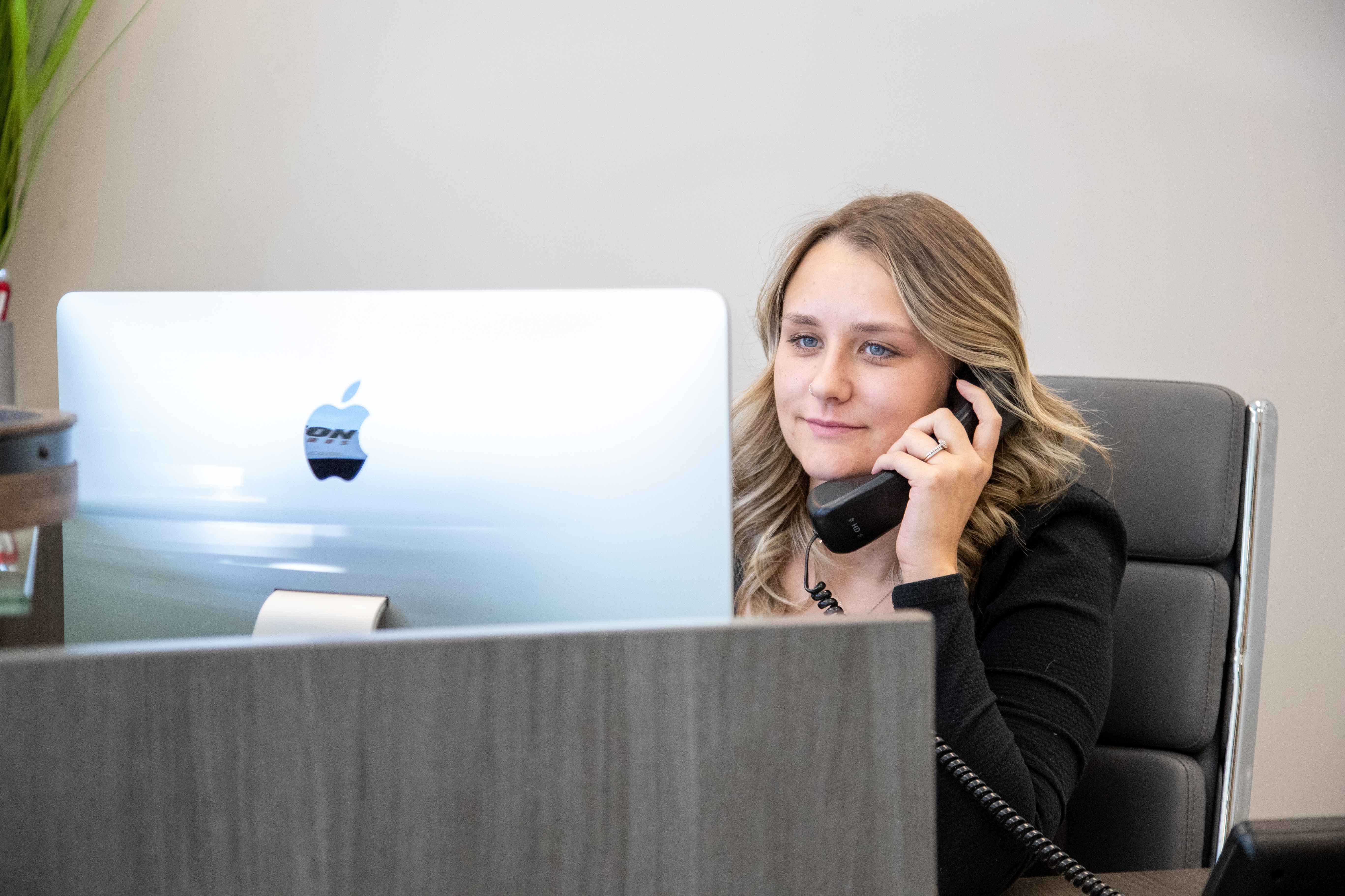 A woman with blonde hair is sitting at a desk, talking on a landline phone and looking at a silver iMac computer screen.
