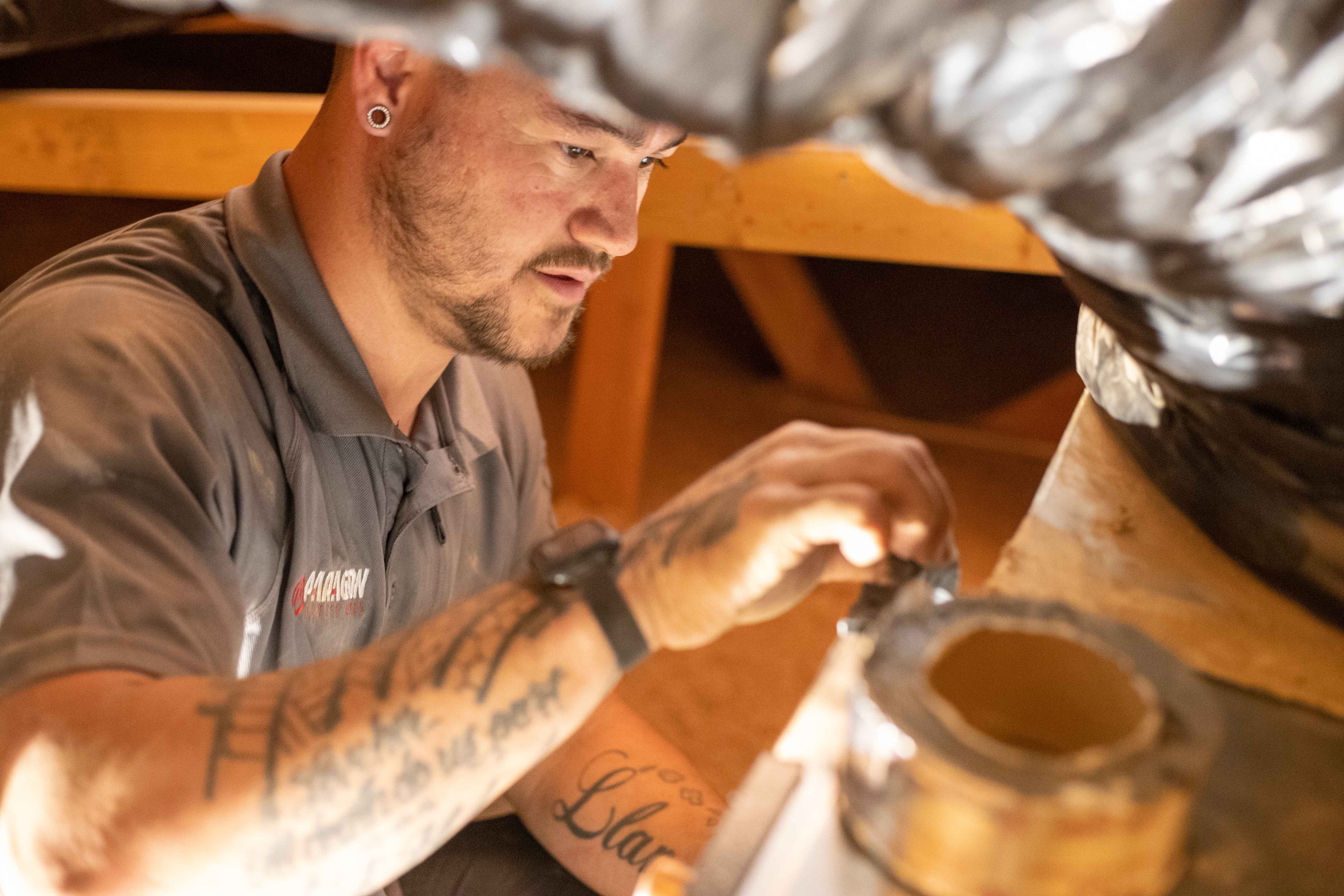Man with tattoos and earring working on heating duct insulation in an attic.