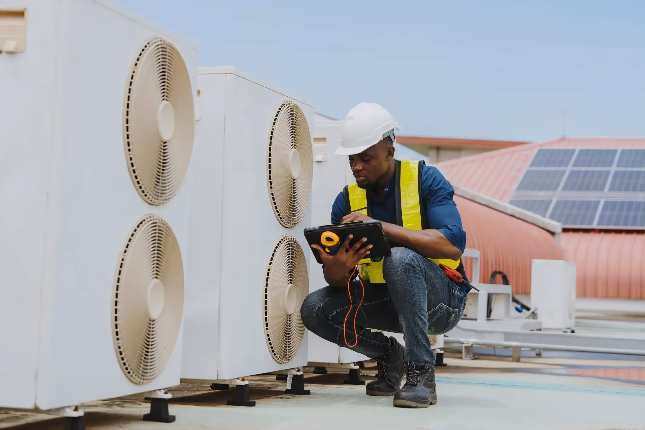 An HVAC technician in a hard hat and safety vest checking an outdoor air conditioning unit on a rooftop with a tablet.