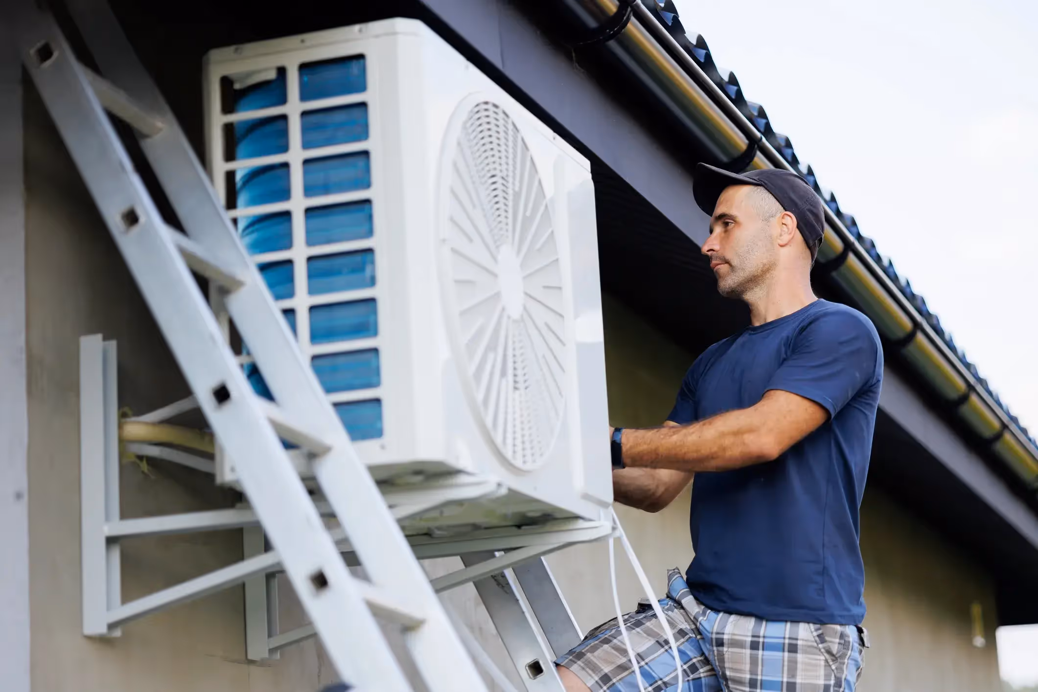 An HVAC technician on a ladder working on an outdoor air conditioning unit attached to a house.
