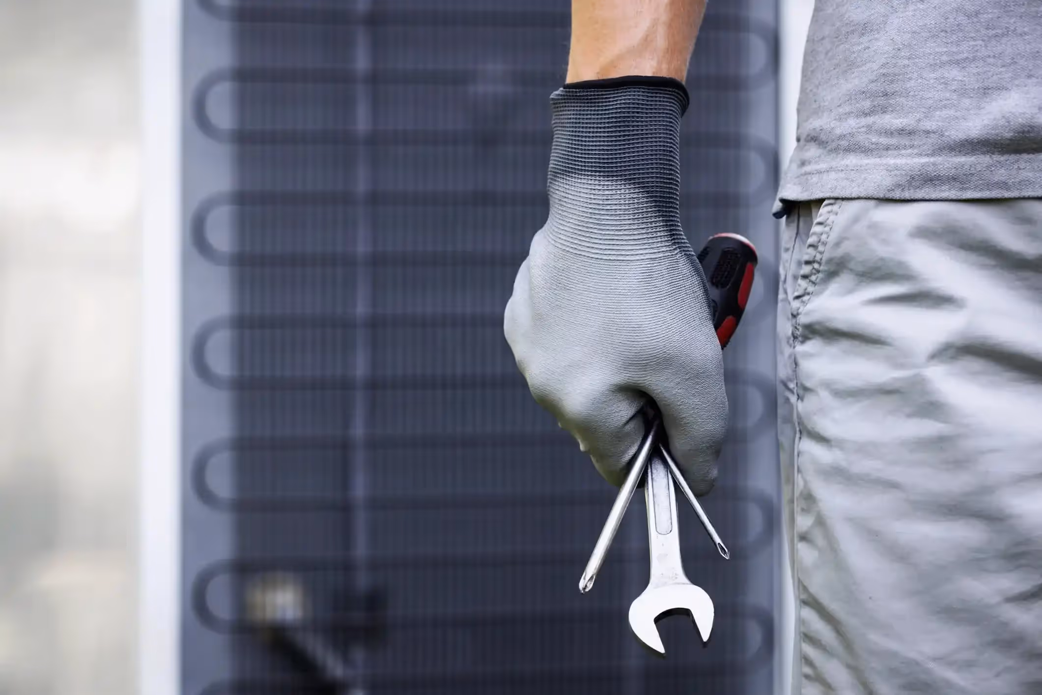 Close-up of a gloved hand holding a wrench and screwdriver in front of an outdoor air conditioning unit.