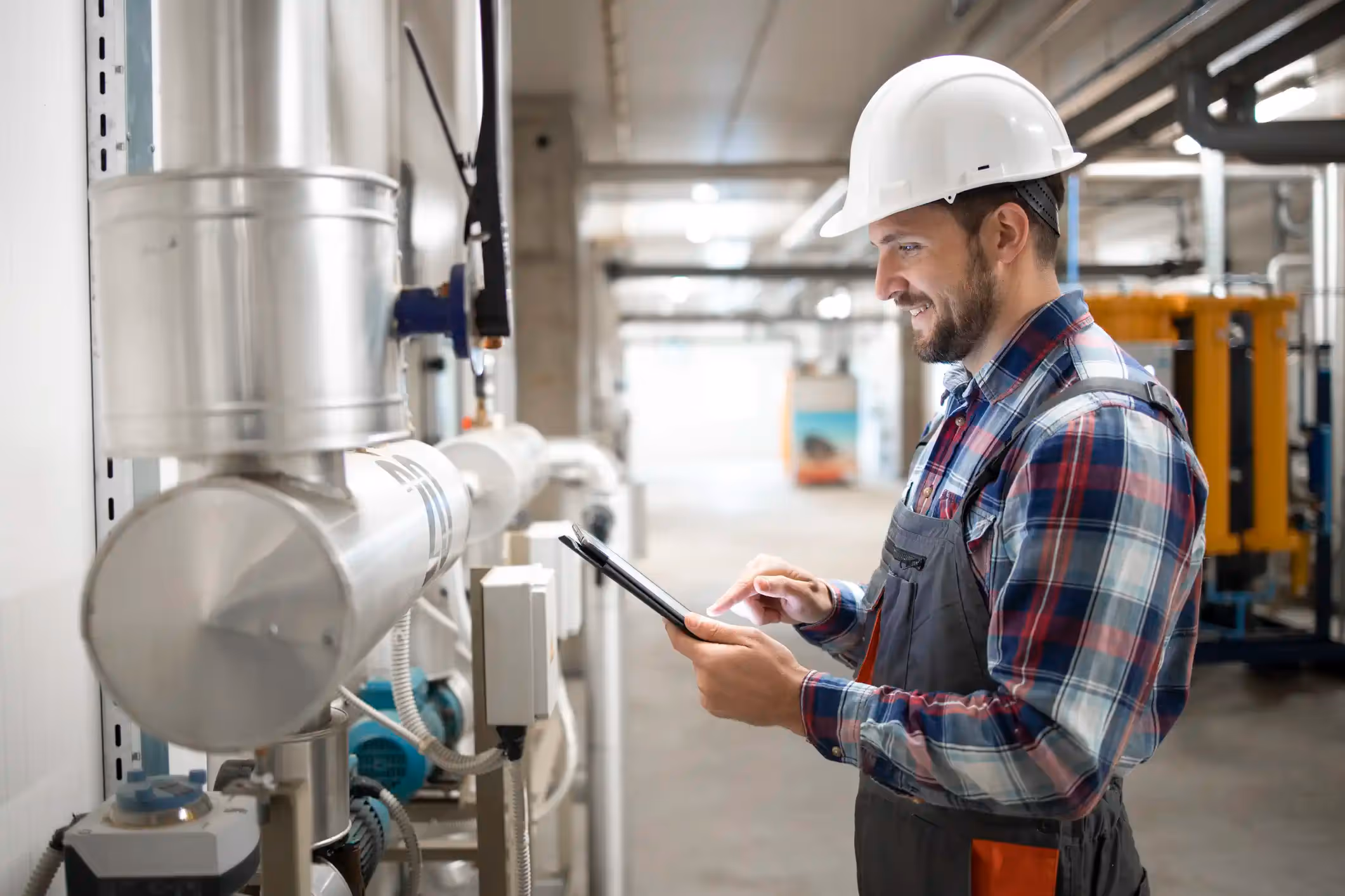  A medium indoor shot of a smiling male engineer or technician in an industrial mechanical room. He is wearing a white hard hat, a plaid button-up shirt (red, white, and blue), and grey overalls. He is standing next to a large, insulated silver pipe and is actively looking at and interacting with a digital tablet he holds in his hands. The background is a brightly lit industrial hallway with equipment, including a prominent yellow machine in the distance, suggesting a large commercial or industrial setting.