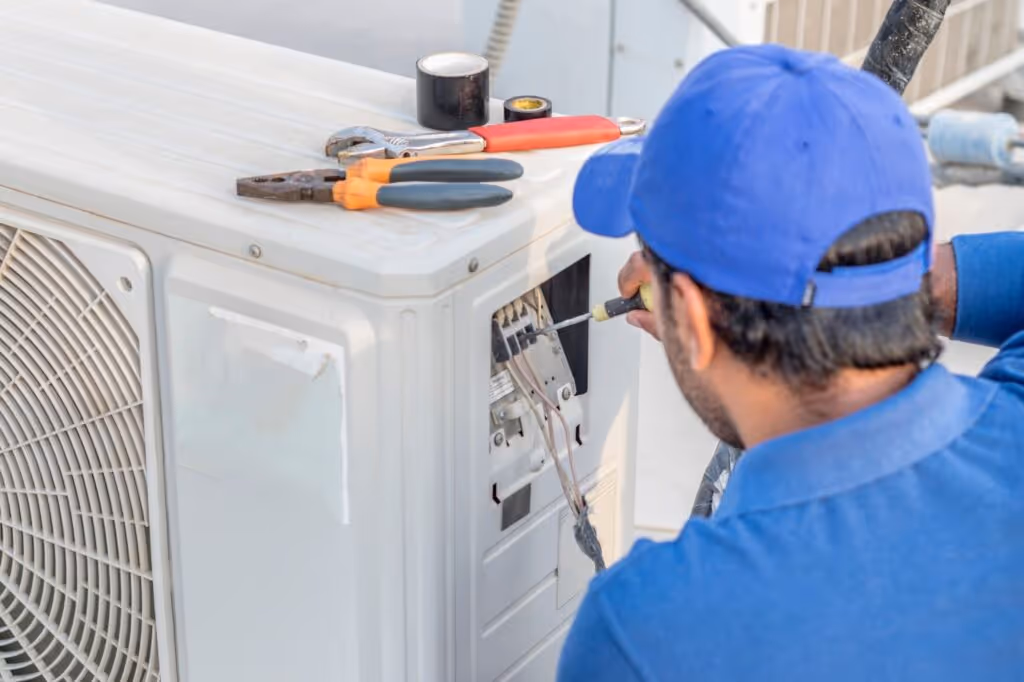 A technician in a blue uniform repairing an outdoor air conditioning unit using a screwdriver.