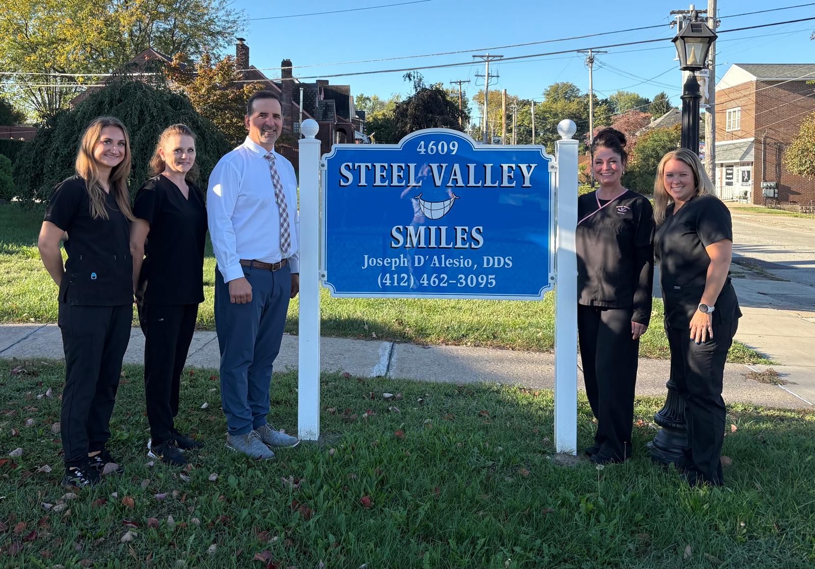 Steel Valley Smiles dental team posing by office sign on sunny day