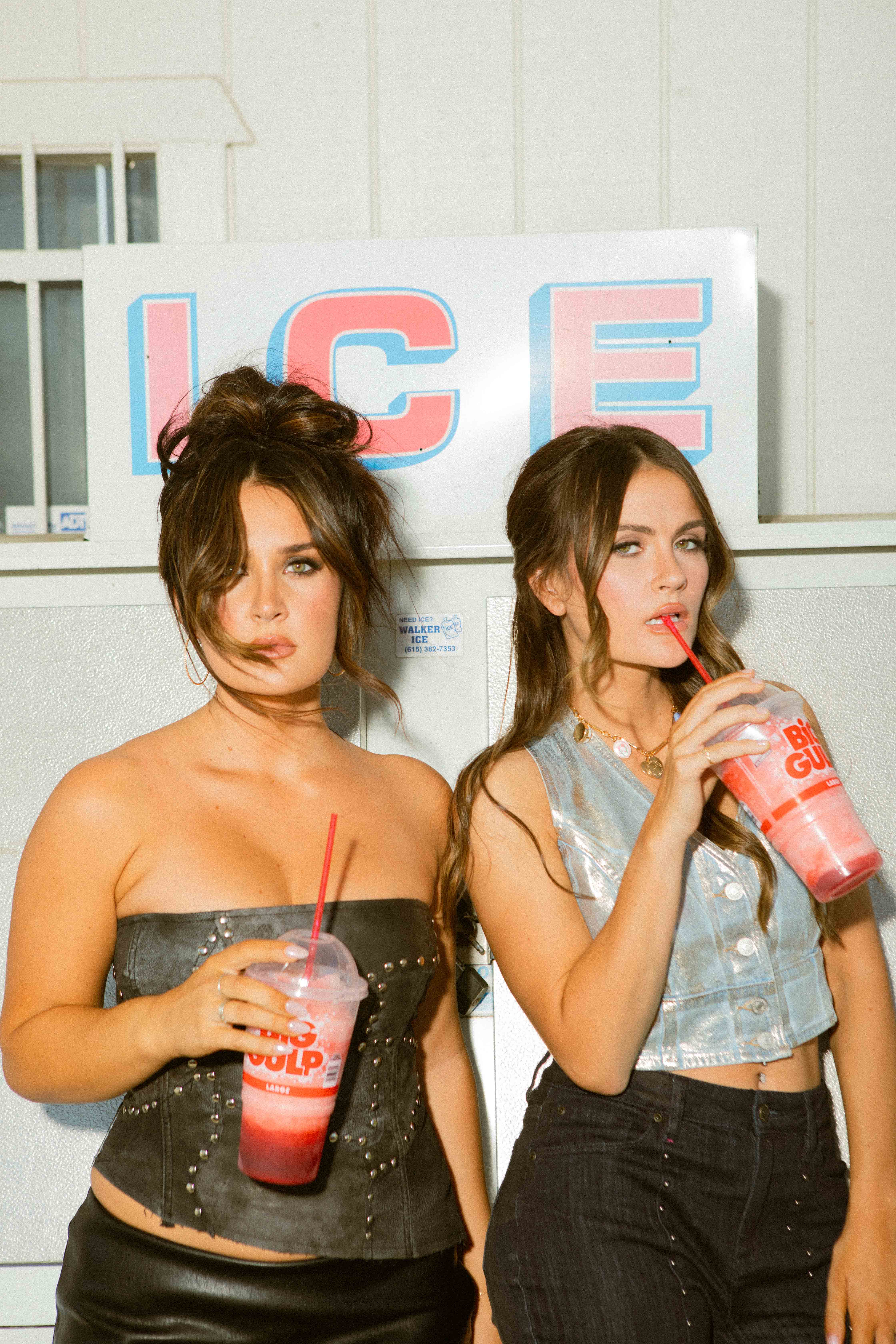 Two women standing in front of an ice vending machine, each holding a large red and white drink with a straw, one woman looks directly at the camera, the other sips her drink.