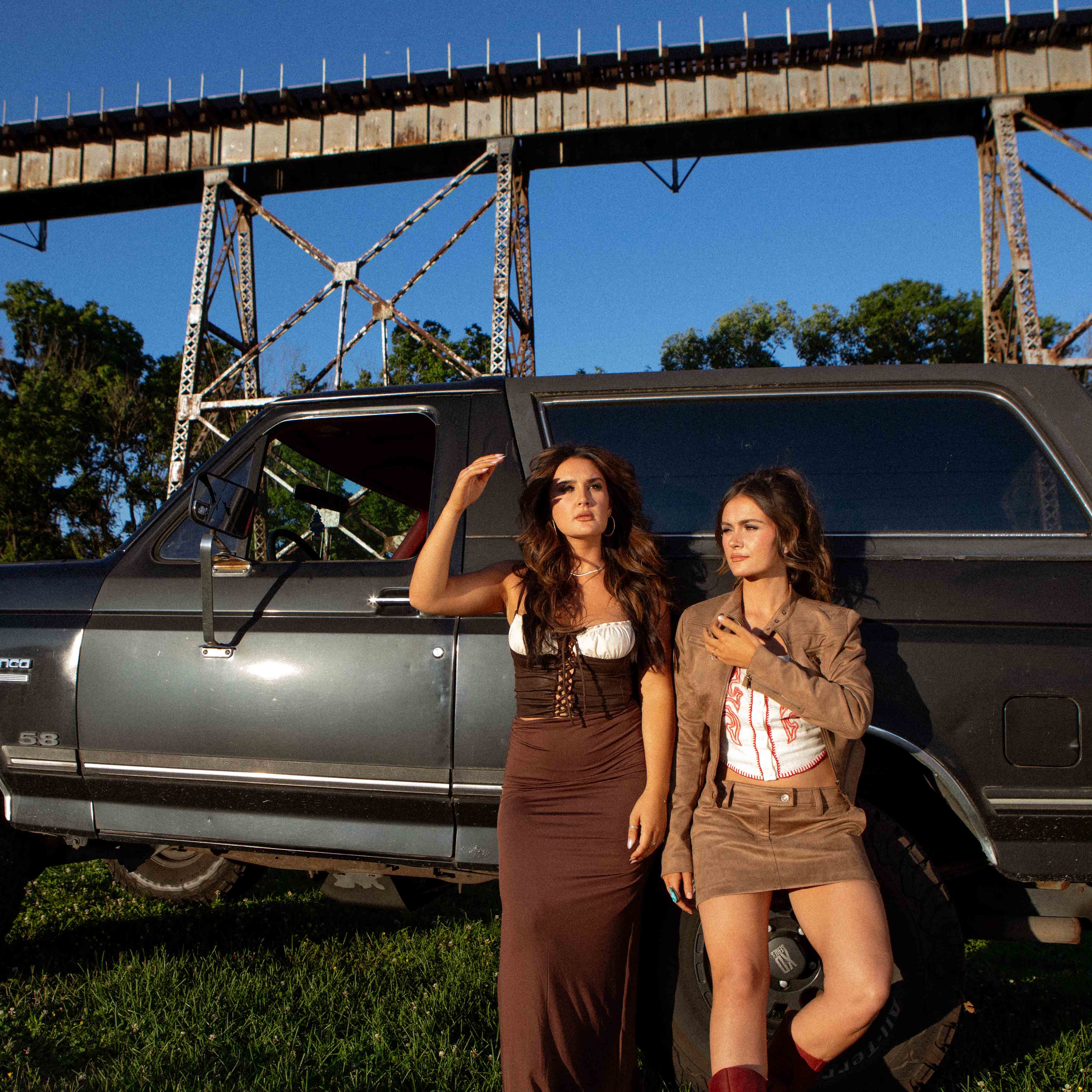 Two women posing beside a dark vintage truck under a rusty railway bridge in a sunny outdoor setting.