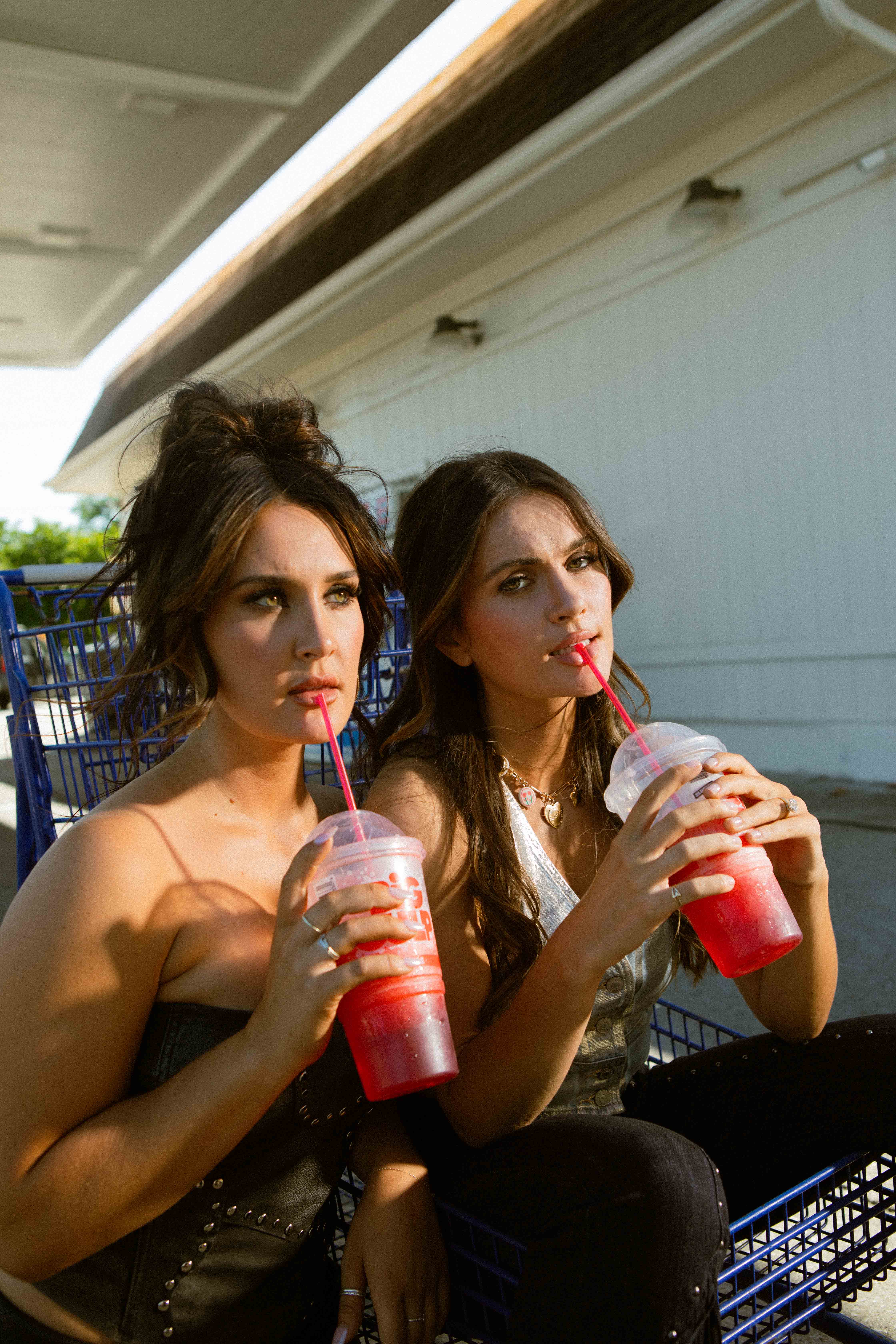Two young women sitting in a shopping cart outdoors, sipping red frozen drinks through straws.