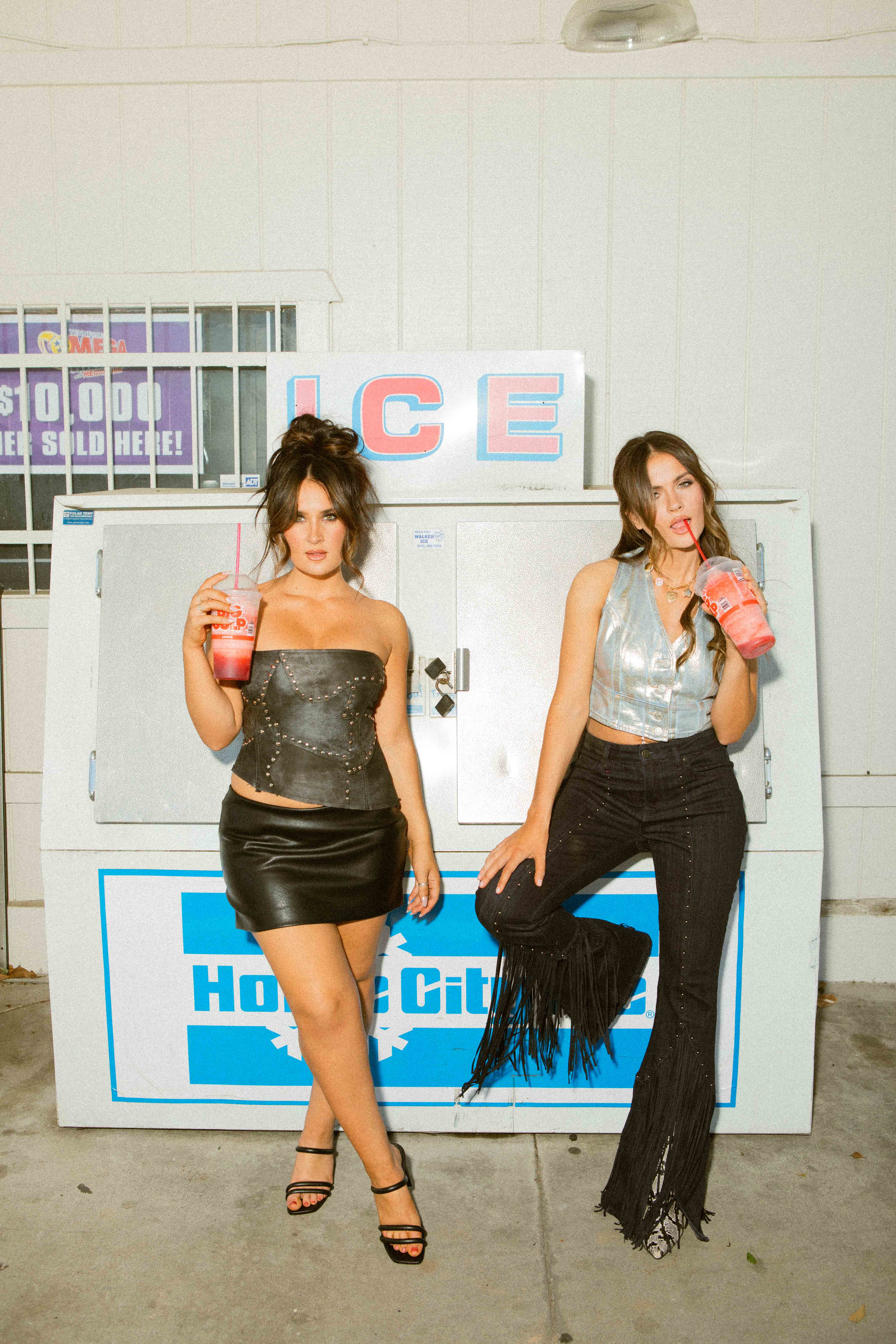Two women standing in front of an ice vending machine, each holding a large red drink with straws; one wears a black leather skirt and strapless top, the other wears black fringed pants and a silver vest.