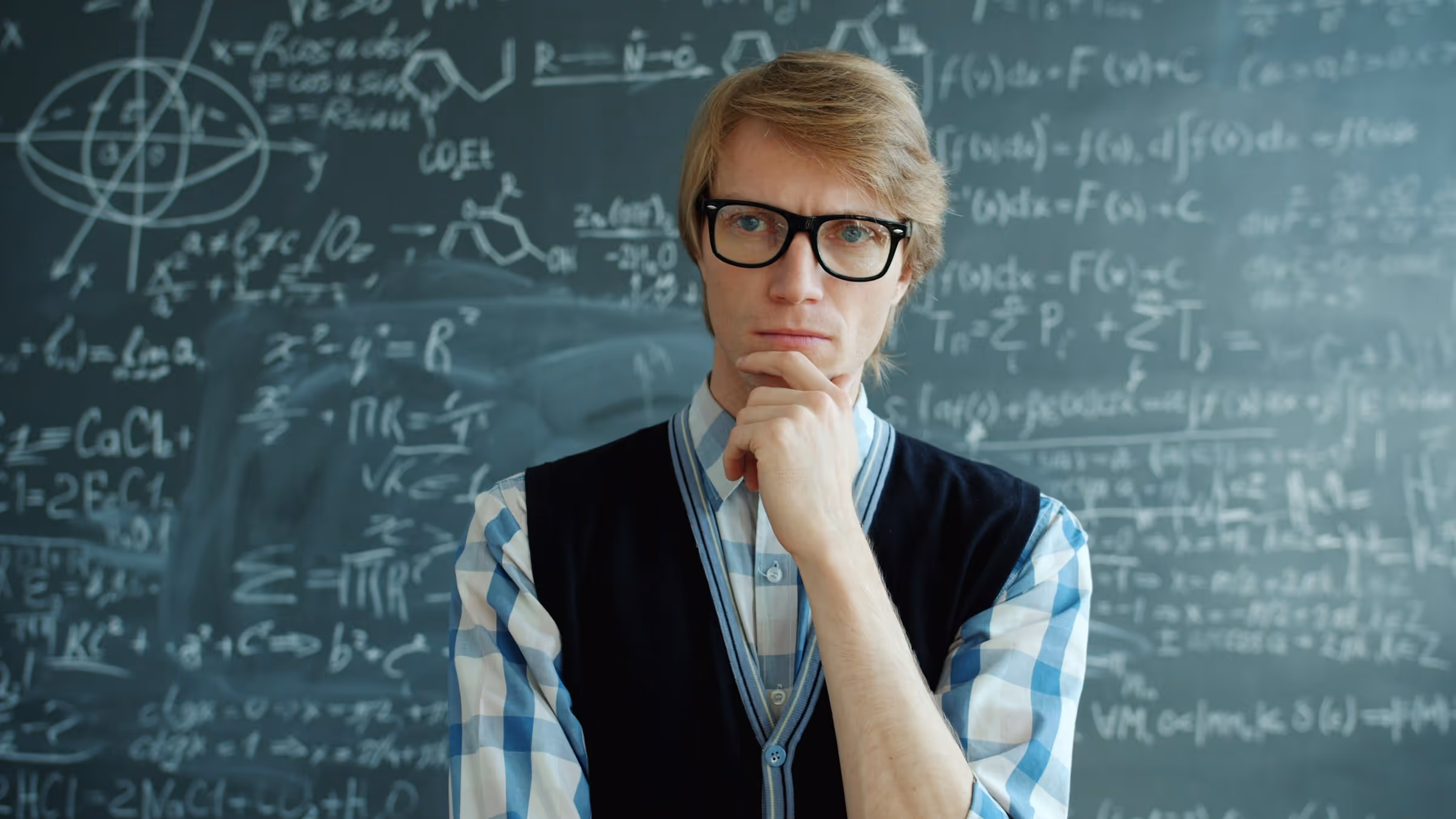 Thoughtful student in glasses standing in front of math equations on chalkboard