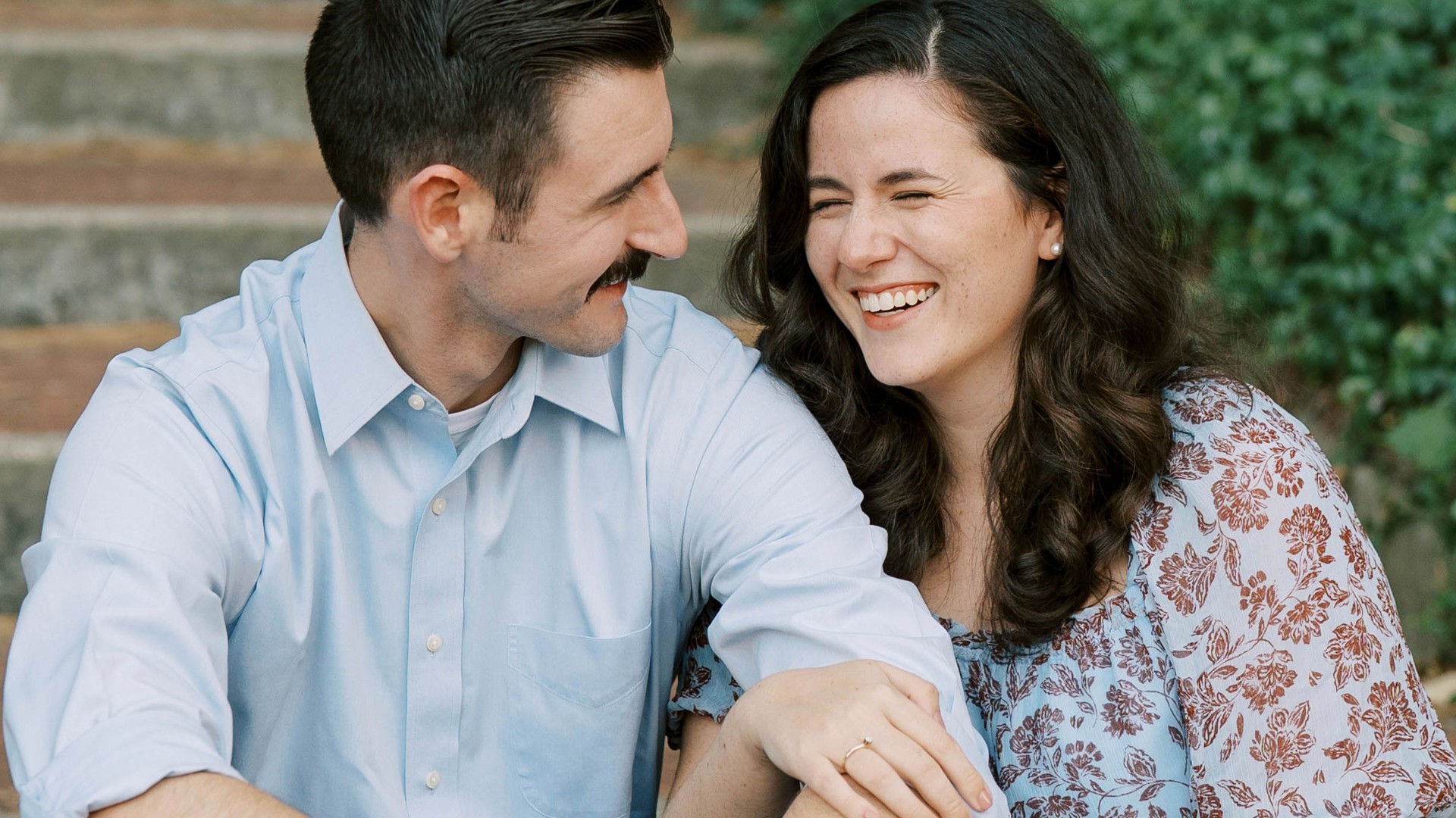 Smiling man in light blue shirt looking at laughing woman with curly hair wearing a floral blouse.