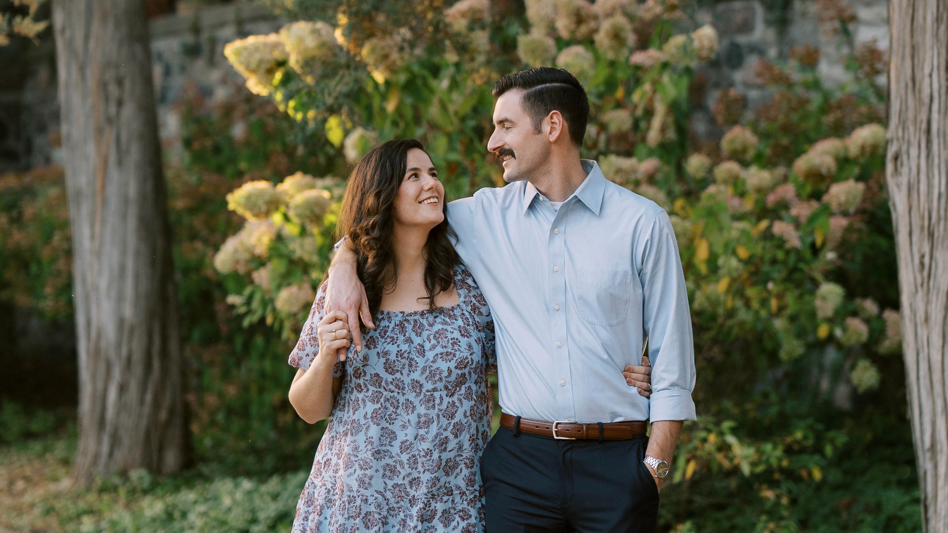 Couple smiling and looking at each other outdoors with greenery and flowers in the background.
