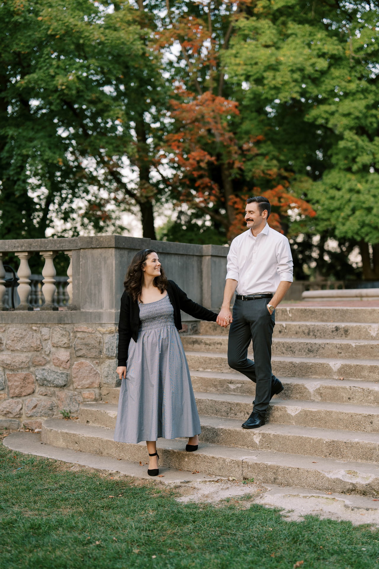 Couple holding hands on stone steps with green and orange trees in the background during autumn.