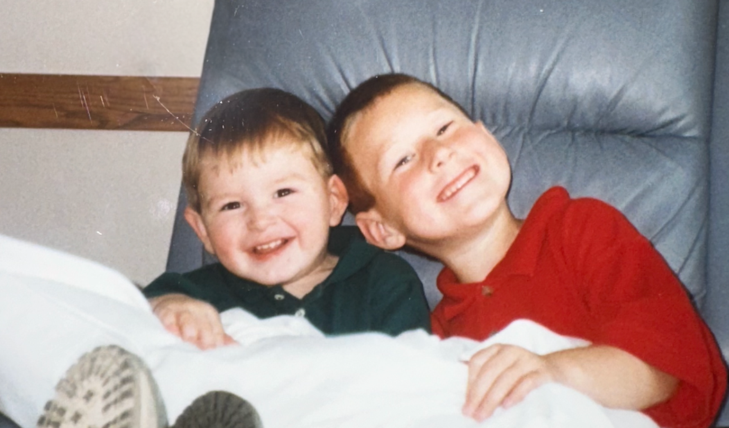 Two young boys smiling and leaning against each other on a blue chair, one in a green shirt and the other in a red shirt.