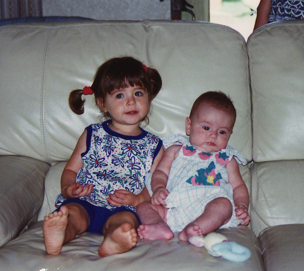 Two young children sitting on a beige leather couch, an older girl with pigtails in a floral top and blue shorts and a baby in a patterned dress.