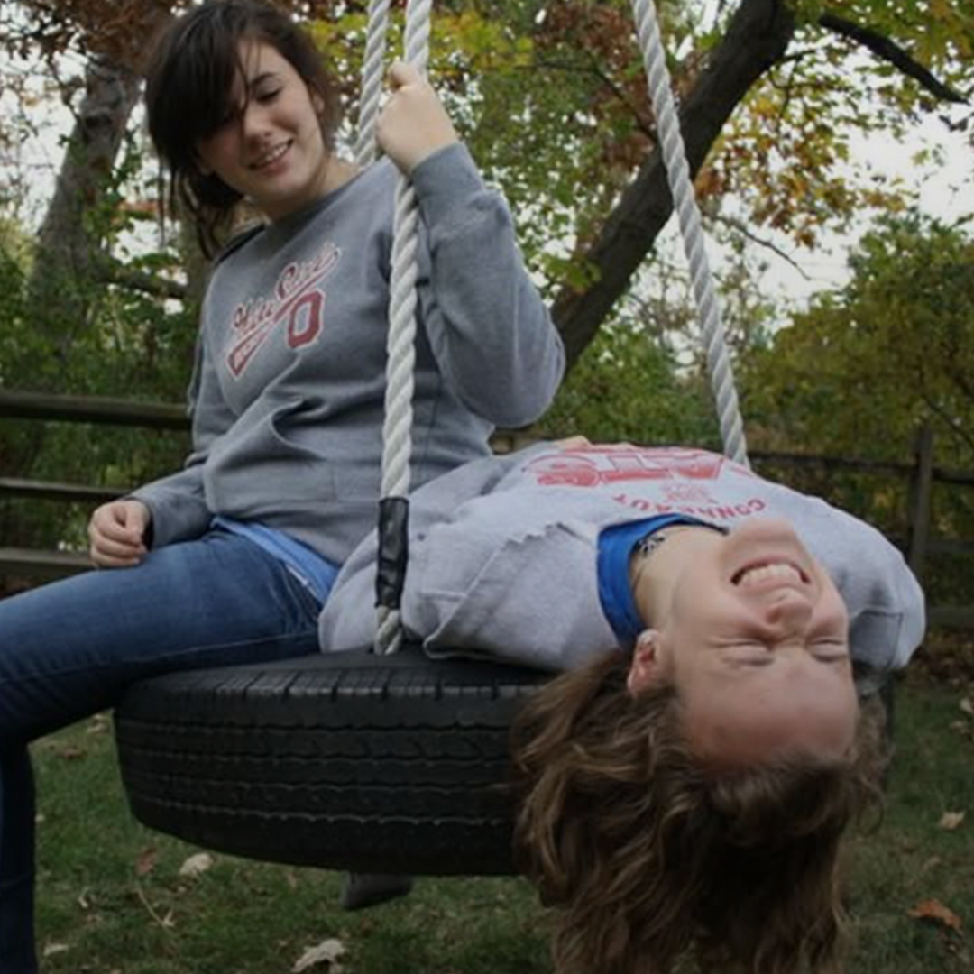 Two girls enjoying on a tire swing, one sitting and smiling, the other laughing with her head tilted back.