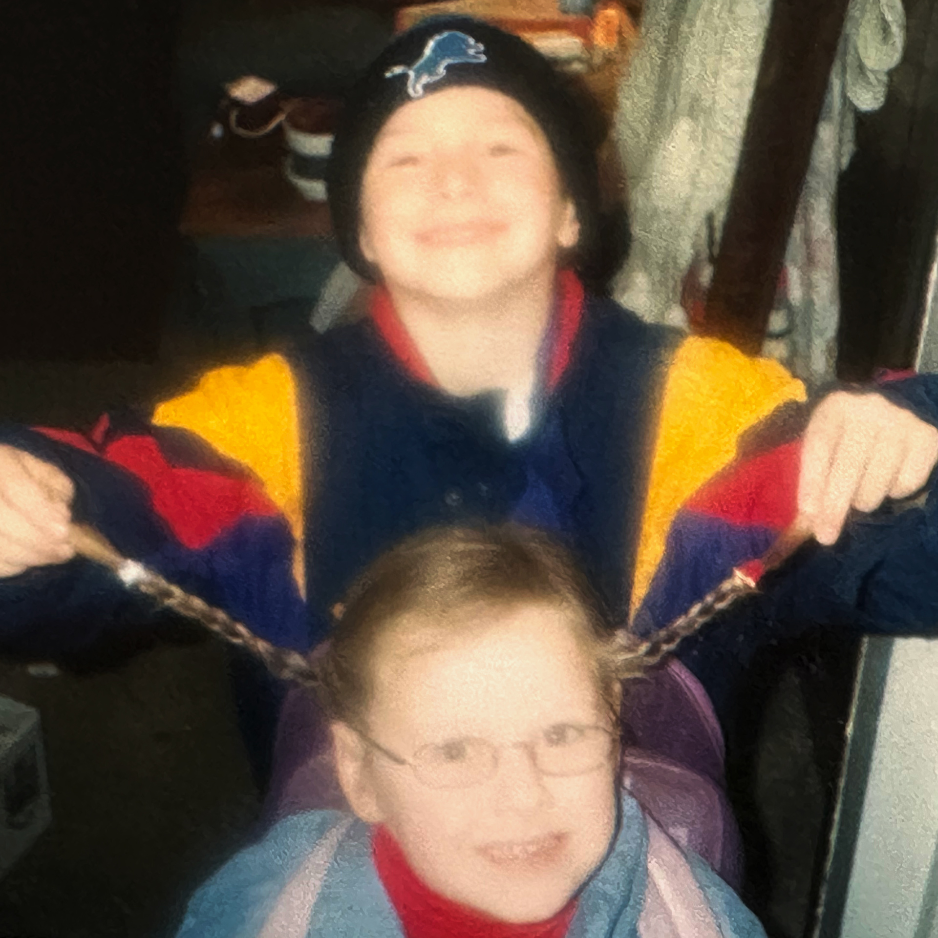 Two children indoors, one wearing glasses smiles in the front while the other behind playfully holds their braided hair out sideways.
