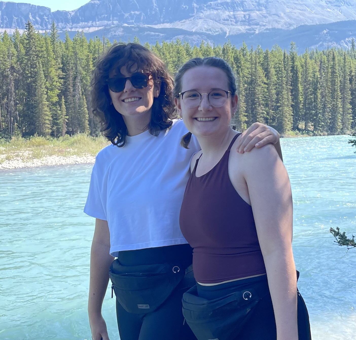 Two smiling women with glasses standing by a river with forest and mountains in the background.
