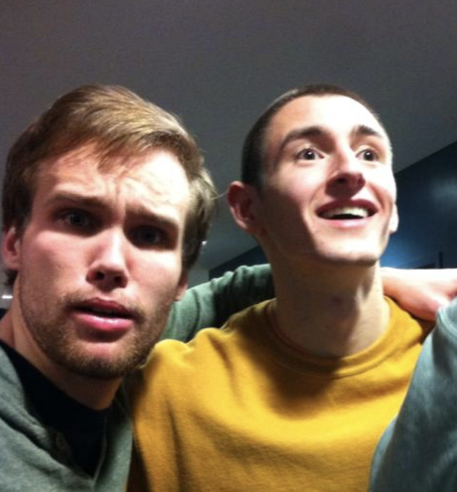 Two young men close together indoors, one wearing a green hoodie looking surprised, the other in a yellow sweater smiling.