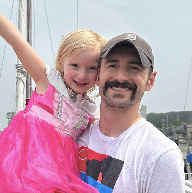 Two young children sitting on a beige leather couch, an older girl with pigtails in a floral top and blue shorts and a baby in a patterned dress.