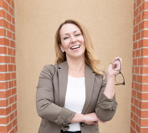 Smiling woman in brown blazer holding glasses standing between two brick pillars with beige background.