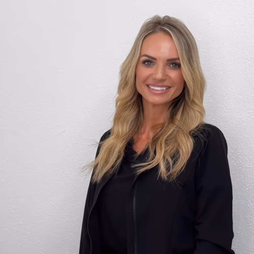 Blonde woman in black outfit standing and smiling in front of a white wall.