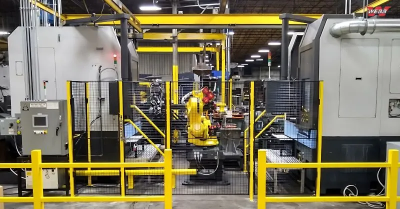 Industrial robotic arm in the center of a factory workstation enclosed by yellow safety barriers and black mesh fencing.