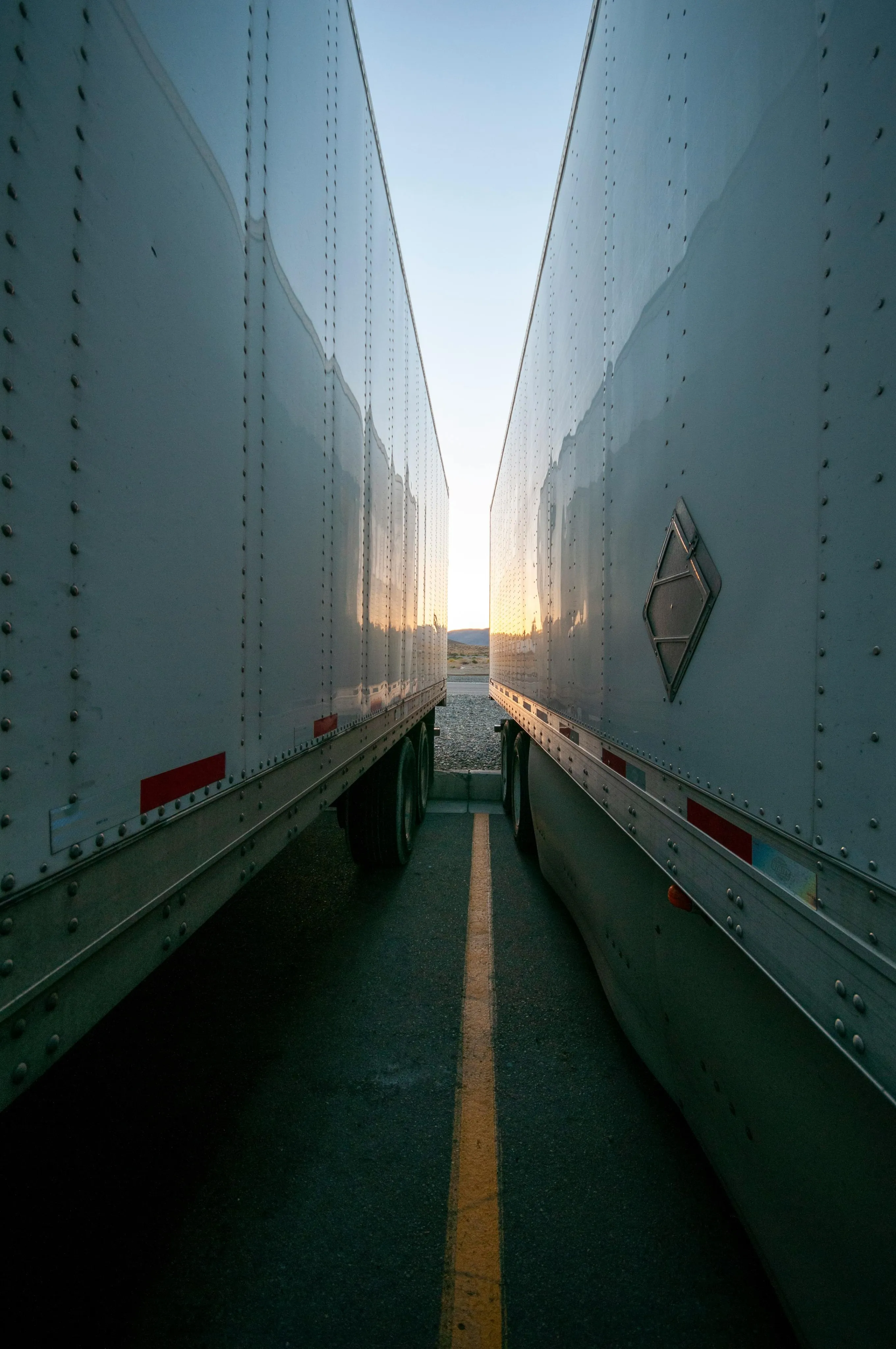 View between two parked white semi-trailer trucks at sunset with a yellow parking line on the asphalt.