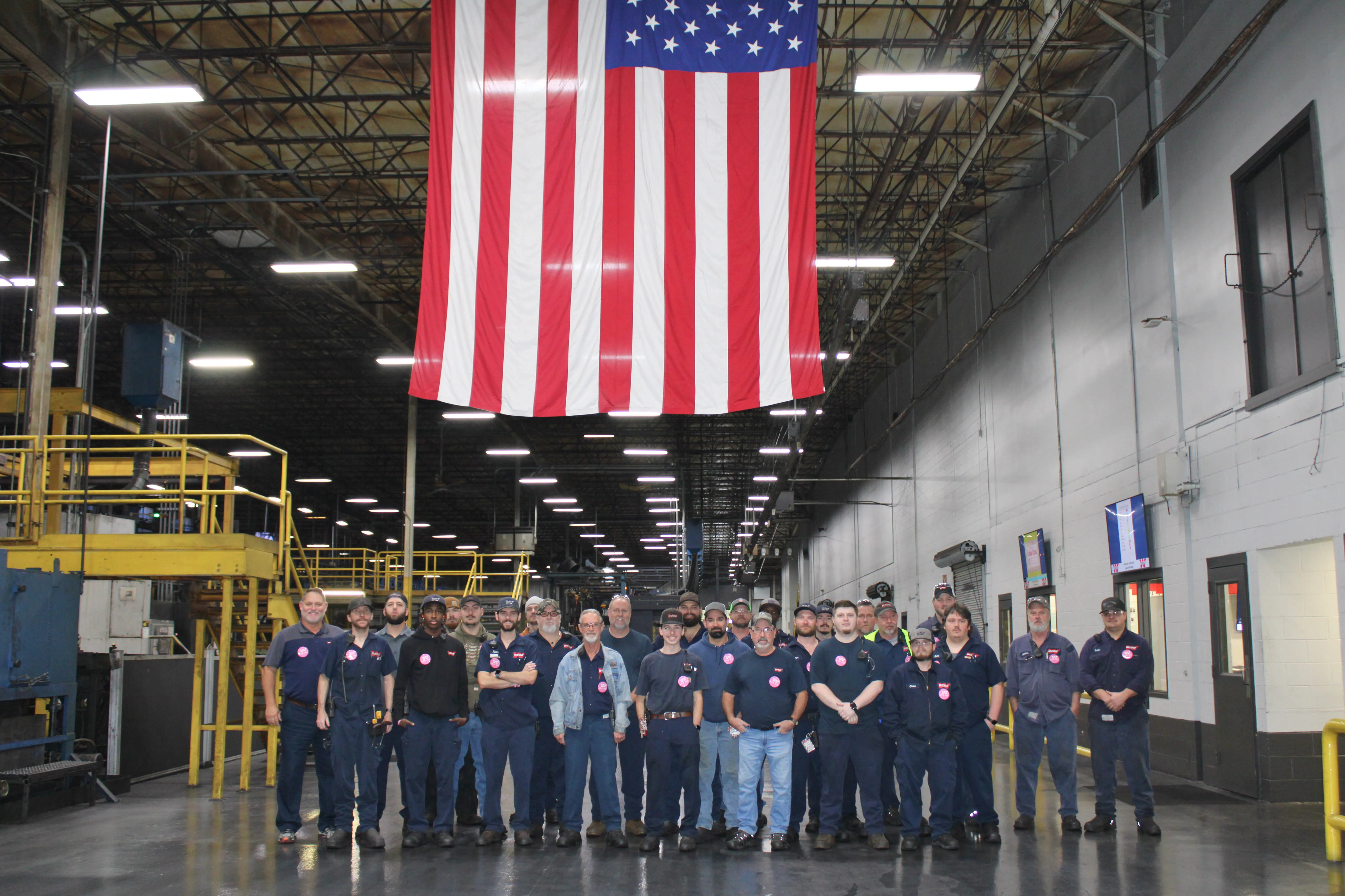 Group of nineteen men standing together inside a large industrial warehouse with an American flag hanging overhead.