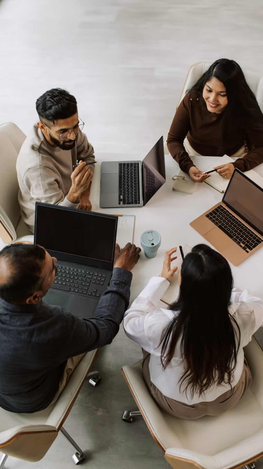 Four diverse colleagues sitting around a white table with laptops, notebooks, and pens, engaged in a discussion.