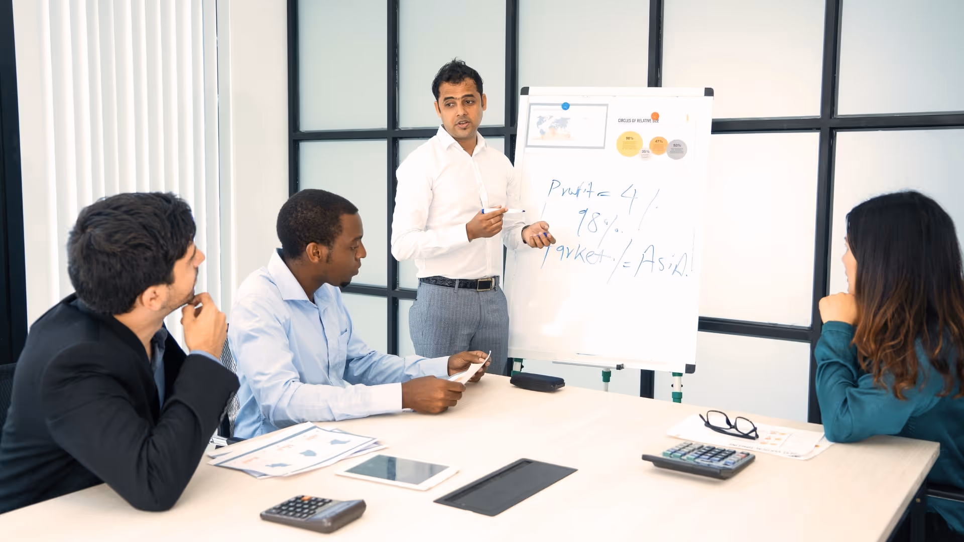 Man in white shirt giving a presentation to two men and a woman seated at a table with documents, calculators, and tablets.