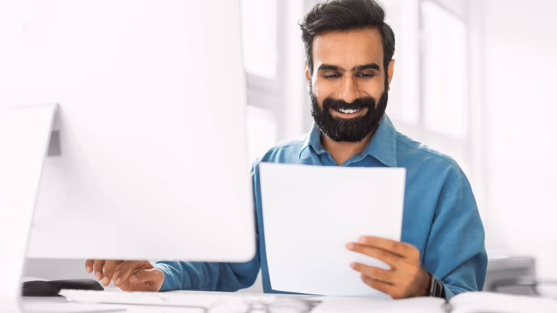 Smiling man with a beard in a blue shirt reading a document at a desk with a computer monitor.